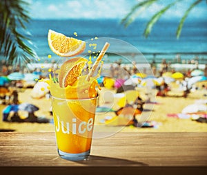 Glass of orange juice with drink splash. Blurred photo of crowded beach at the background. Summer vacation concept