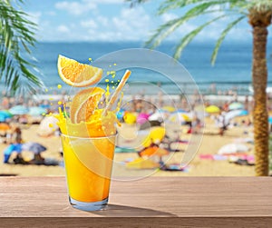 Glass of orange juice with drink splash. Blurred photo of crowded beach at the background. Summer vacation concept