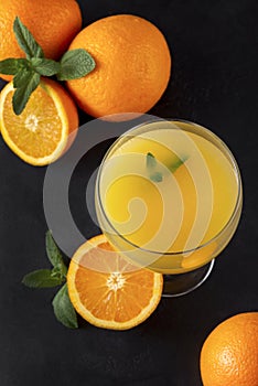 Glass of orange juice with mint, fresh whole and half oranges on a black background, citrus drink top view