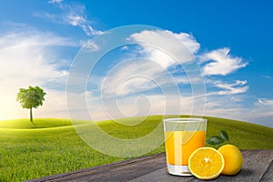 A glass of orange juice and oranges on old wooden floor beside green field on slope and tree with blue sky and clouds background