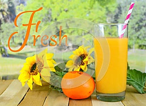 A glass of orange juice sitting on a wooden table with sunflowers and a single orange with a blurred background of a tree lined la