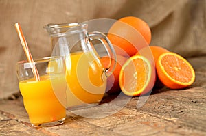 Glass with orange juice and straw, jug with fresh juice and pile of oranges in the background on wooden table