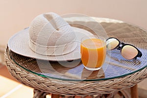 A glass of orange juice, sunglasses and a grey hat are on a wicker table illuminated by sunlight.