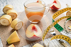 A glass of peach juice on a sackcloth on a white wooden table with slices of fruit and a fork and a centimeter