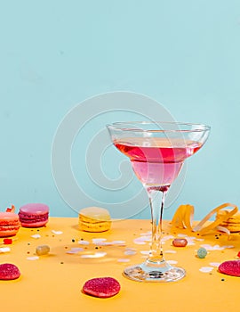 Glass of pink cocktail with heart candies and macaroons on yellow table. Birthday party or Valentines day concept
