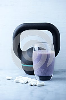 Glass of Protein Shake with milk and blueberries. Creatine capsules and black sporting kettlebell in background. Sport nutrition.