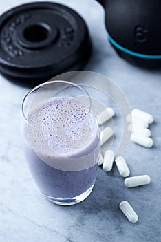 Glass of Protein Shake with milk and blueberries. Creatine capsules, black sporting kettlebell and plates in background. Sport nut