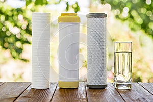 A glass of purified water and filter cartridges on wooden table on green natural background