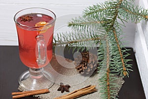 A glass of red drink stands on a piece of linen. A slice of dried orange and anise floats in it. Nearby are cinnamon sticks and