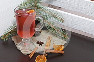 A glass of red drink stands on a piece of linen. A slice of dried orange and anise floats in it. Nearby are cinnamon sticks and