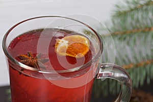 A glass of red drink stands on a piece of linen. A slice of dried orange and anise floats in it. Nearby are cinnamon sticks and