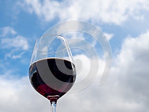 A glass of red wine against a blue sky with white clouds.