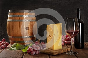 A glass of red wine with a bottle, a wine barrel, a large piece of cheesse, grapes, and vine leaves