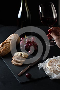 Glass of red wine with a bottle, cluster of grapes, women hand placing brie cheese to rustick piece of bread with antique knife.