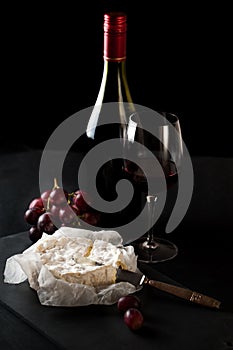 Glass of red wine with a bottle, cluster of grapes, women hand placing brie cheese to rustick piece of bread with antique knife.