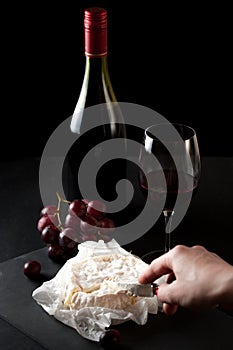 Glass of red wine with a bottle, cluster of grapes, women hand placing brie cheese to rustick piece of bread with antique knife.