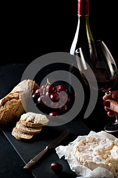 Glass of red wine with a bottle, cluster of grapes, women hand placing brie cheese to rustick piece of bread with antique knife.