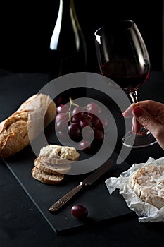 Glass of red wine with a bottle, cluster of grapes, women hand placing brie cheese to rustick piece of bread with antique knife.