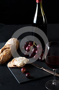 Glass of red wine with a bottle, cluster of grapes, women hand placing brie cheese to rustick piece of bread with antique knife.