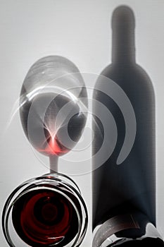 Glass of red wine and bottle with shadows and reflections on a white textured surface.