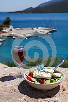 A glass of red wine and bowl of greek salad with greek flag on by the sea view, summer greek holidays concept.