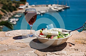 A glass of red wine and bowl of greek salad with greek flag on by the sea view, summer greek holidays concept.