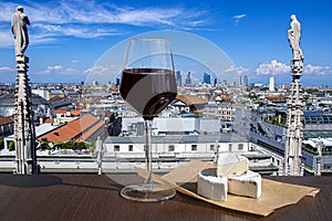Glass of red wine with brie cheese against view from above of Milan, Italy