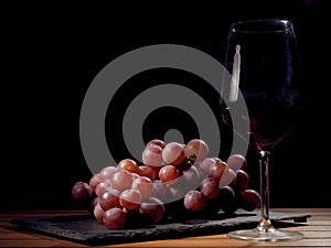 Glass of red wine and cluster of red fresh organic grapes on a black slate plate and wooden table.