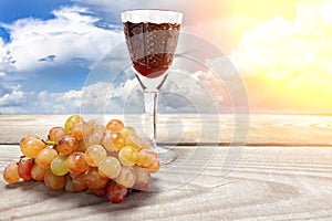 A glass of red wine with grapes on a wooden table against a background of clouds
