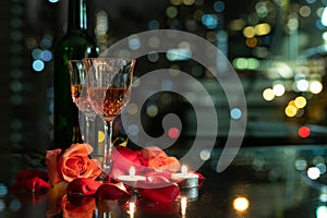 Glass red wine, roses, petals and candles on table with reflection, twinkling blurred city night lights in background, romantic