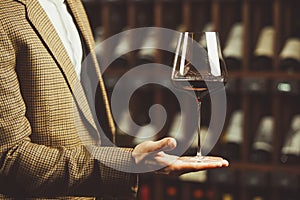 Glass of red wine in the sommelier hand on the cellar with bottles backgrounds.