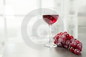 A glass of red wine stands on a table next to a bunch of red grapes. White kitchen with a large panoramic window on the background