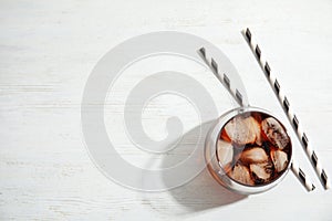 Glass of refreshing cola with ice cubes on white wooden background, top view.