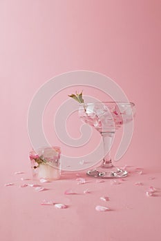 Glass with rose petals and an ice cube with a flower next to it on pink background