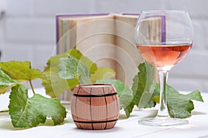 Glass with rose wine and a wooden barrel with a branch and leaves of grapes and a book on a white wooden table.