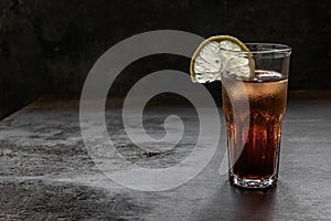 A glass of rum with cola full of ice with a slice of lemon on a dark brown old bar table in grunge style. Copy space