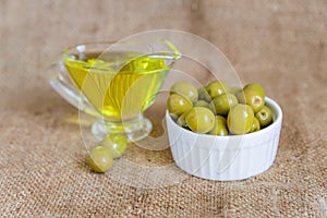 Glass sauceboat with extra virgin olive oil and fresh green olives in white ceramic bowl on  burlap cloth background