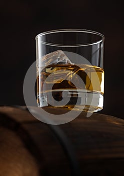 Glass of single malt whiskey with ice cubaes on top of wooden barrel on black