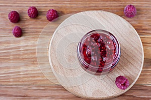 Glass small saucer with raspberry jam with fresh raspberries close up on wooden background.Copy space