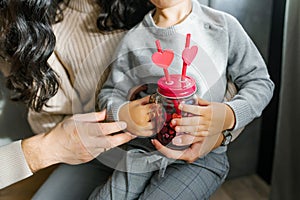 A glass smoothie mug with two tubes with hearts in the hands of a young son. Valentine`s Day in the family