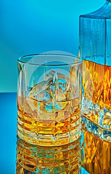 A glass and square crystal decanter with scotch whiskey or brandy in the background on a blue gradient background with reflection