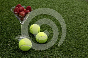 Glass of strawberries and tennis balls on grass.