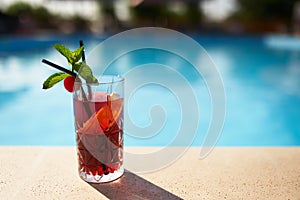 Glass of strawberry cocktail decorated with mint and cherry on swimming pool deck. Refreshing alcohol drink on water