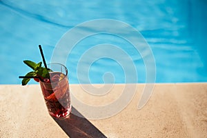 Glass of strawberry cocktail decorated with mint and cherry on swimming pool deck. Refreshing alcohol drink on water