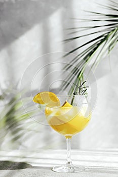 A glass with a summer cocktail with an orange and a sprig of rosemary under the branches of palm trees and the bright sun. Hard