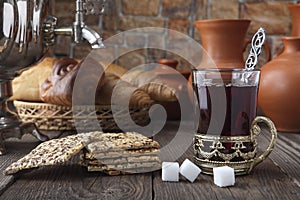 A glass of tea with a cracker and rolls near the samovar and ceramic dishes on an old table. Retro stylized photo.