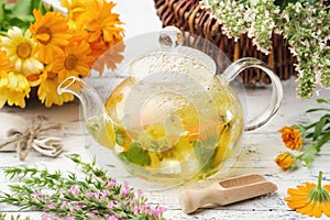Glass tea kettle of healthy herbal tea, hyssop, calendula, balm mint medicinal herbs on white wooden table.