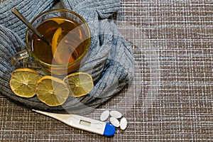 A glass of tea with lemon wrapped in a wool gray scarf. On the table lies a thermometer and pills.