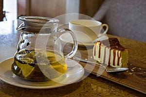 Glass teapot with green tea, mint and lemon, cup of tea, small teaspoon and half-eaten piece of cheesecake on table in cafe