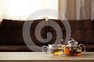 Glass teapot and thermo cup with green tea and dried apricots on wooden table against defocused sofa with pillows. Front view.